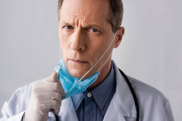 mature doctor in latex glove touching blue medical mask on grey  - Фото, изображение