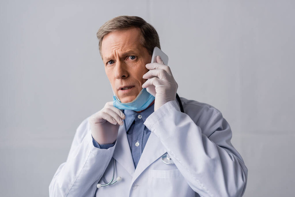 stressed and mature doctor talking on smartphone on grey  - Foto, Imagem