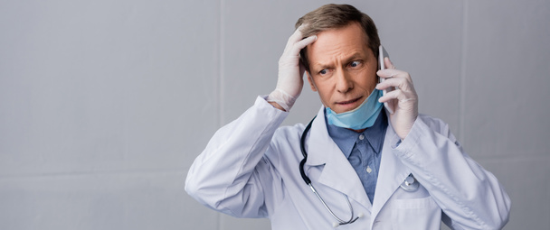 панорамний знімок стресового лікаря середнього віку, який говорить на смартфоні на сірому
  - Фото, зображення