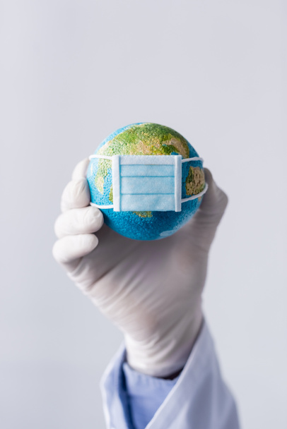 Ausgeschnittene Ansicht eines Arztes mittleren Alters im Latex-Handschuh mit kleinem Globus in medizinischer Maske isoliert auf grau - Foto, Bild