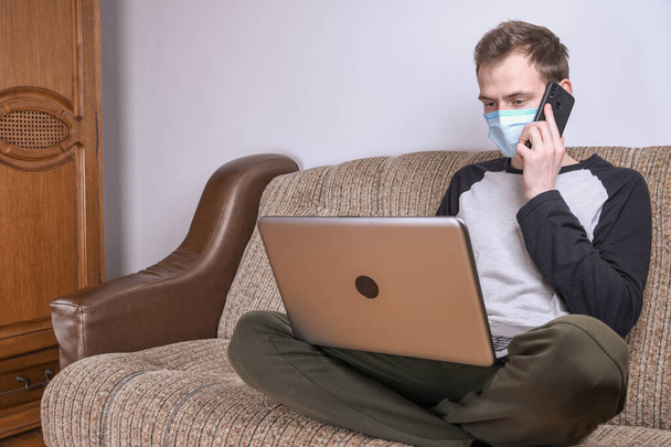 Hombre joven con máscara médica que trabaja en casa en la habitación en el sofá utilizando un ordenador portátil. Cuarentena, autoaislamiento, protección contra el coronavirus. Vacaciones
. - Foto, Imagen
