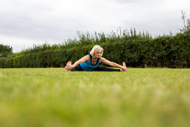 Uma mulher de meia-idade praticando ioga descalça lá fora em um parque gramado. Ela está usando um colete azul brilhante e leggings pretos. O estilo de ioga que ela está fazendo é Hatha Yoga - Foto, Imagem