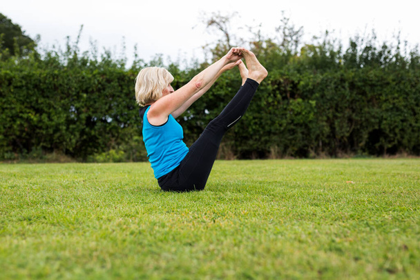 Una mujer de mediana edad practicando yoga descalza afuera en un parque cubierto de hierba. Lleva un chaleco azul brillante y polainas negras. El estilo de yoga que está haciendo es Hatha Yoga - Foto, imagen