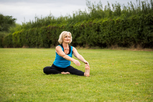 Una mujer de mediana edad practicando yoga descalza afuera en un parque cubierto de hierba. Lleva un chaleco azul brillante y polainas negras. El estilo de yoga que está haciendo es Hatha Yoga - Foto, imagen