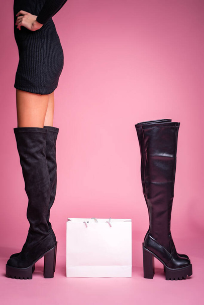 Το κορίτσι στέκεται με μαύρες μπότες στα πόδια της σε ροζ φόντο. Στη μέση υπάρχει μια τσάντα από το κατάστημα. Υπάρχει ένα άλλο ζευγάρι μπότες Hessian στο πλάι. Μοντέλο στις μπότες ώρα πάνω από το γόνατο. γυρίσματα στούντιο. Λεπτά πόδια. μοντέρνα μπότα - Φωτογραφία, εικόνα