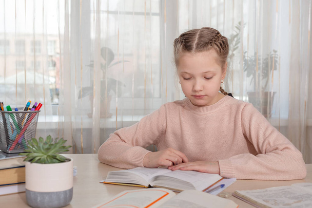 Schoolmeisje studeert thuis. Een meisje dat een boek leest aan tafel. Afstandsonderwijs, thuisonderwijs. Zelfisolatieconcept - Afbeelding - Foto, afbeelding