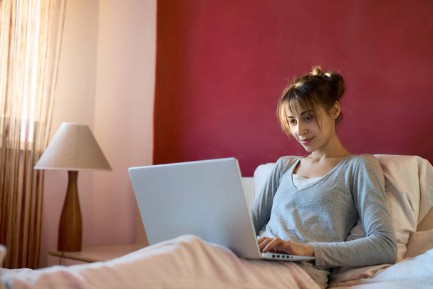 skilled female freelancer working on laptop from her home bed, bright red wall on background. Concept remote work, freelance, internet business, - Foto, Bild