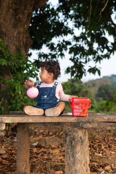Margao, Goa / India - Április 2 2020: Játékos csinos indiai lány gyermek / csecsemő visel sapkát, olvas egy könyvet, és játszik, amelynek mezők és naplemente a háttérben. A kölyök vidám kifejezéseket ad. - Fotó, kép