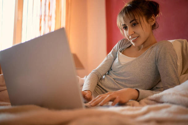 mujer sonriente relajarse en la cama en casa y charlar con amigos en las redes sociales en el ordenador portátil, el uso de Internet velocidad inalámbrica. Mujer comunicándose a través de chat de vídeo en línea, escribiendo en el teclado del ordenador portátil
. - Foto, imagen