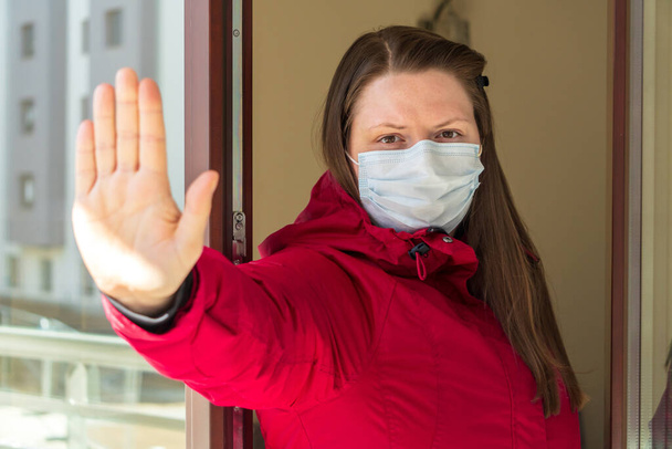 Menina ou jovem mulher em uma máscara médica que está sendo colocado em quarentena em casa durante a pandemia de coronavírus. Auto-isolamento, vista através de vidro, andando na varanda de um apartamento na cidade
 - Foto, Imagem