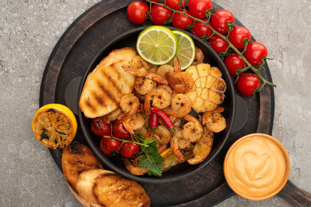 top view of fried shrimps with grilled toasts, vegetables, cherry tomatoes and lime on wooden board with sauce on grey concrete background - Fotografie, Obrázek