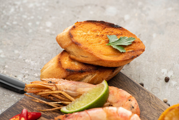 close up view of fried shrimps with grilled toasts and lime on wooden board on grey concrete background - Fotografie, Obrázek