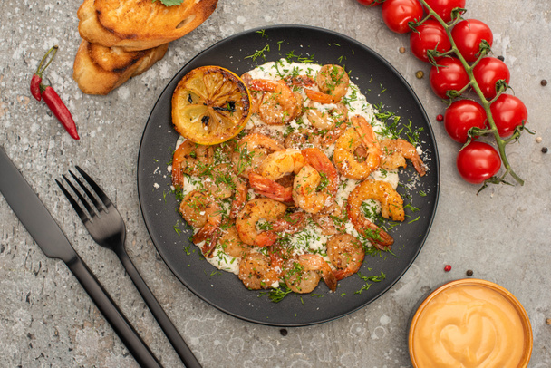 top view of fried shrimps in sauce with dill and lemon near cutlery, cherry tomatoes and toasts on grey concrete background - Fotografie, Obrázek
