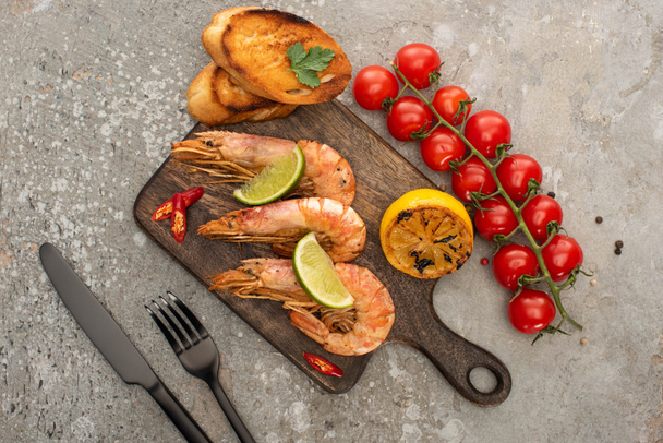 top view of fried shrimps with grilled toasts, lemon and lime near cherry tomatoes on wooden board near cutlery on grey concrete background - Photo, Image