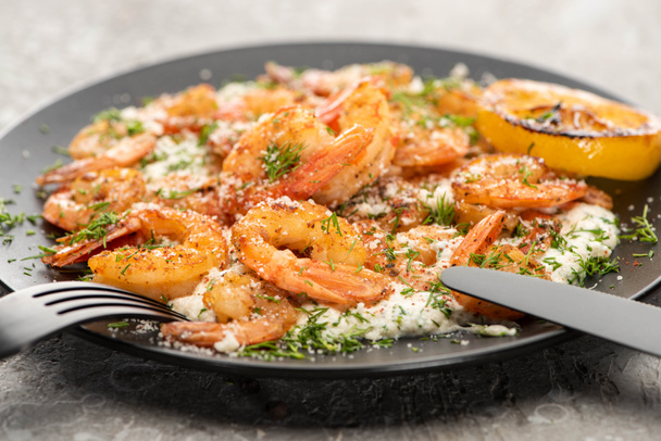 selective focus of fried shrimps in sauce with dill and lemon in black plate on grey concrete background with cutlery - Foto, Imagen