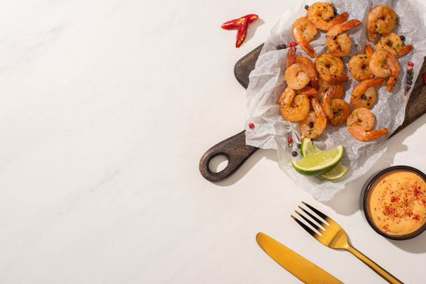 top view of fried shrimps on parchment paper on wooden board with cutlery, chili pepper, sauce and lime on white background - Photo, Image