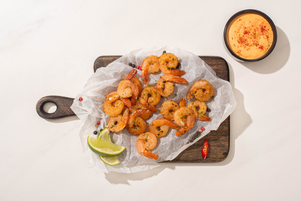 top view of fried shrimps on parchment paper on wooden board with chili pepper, sauce and lime on white background - Фото, зображення