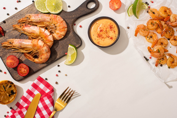 top view of fried shrimps on parchment paper and wooden board with cutlery, plaid napkin, vegetables, sauce and lime on white background - Photo, Image