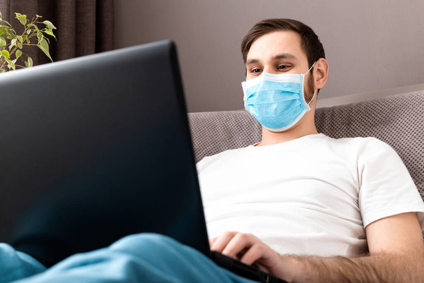 Young caucasian man working from home office wearing protective mask using laptop and internet. Coronavirus covid 19 quarantine. Remote work, freelancer, cozy home office workplace on sofa. - Foto, afbeelding