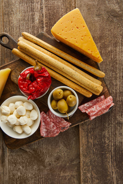 Bovenaanzicht van schalen met antipasto ingrediënten in de buurt van broodstokjes, salami plakjes en kaas op planken op houten ondergrond - Foto, afbeelding