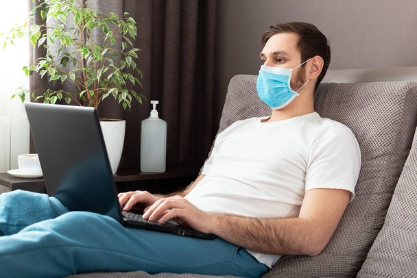 Young caucasian man working from home wearing protective mask using laptop and internet. Cozy Home office, workplace on sofa during coronavirus pandemic, covid 19 quarantine. Remote work, freelancer. - Photo, image