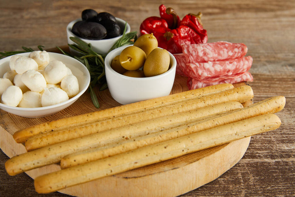 Round board with breadsticks, salami slices, greenery and bowls with antipasto ingredients on wooden background  - Photo, Image