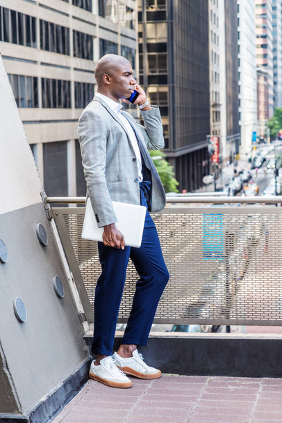 Jovem afro-americano vestindo blazer cinza estampado, calças azuis, tênis brancos, segurando computador portátil, de pé por trilhos na varanda, de frente para a rua com prédios altos, falando no celular
. - Foto, Imagem