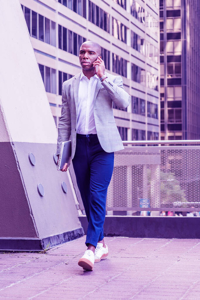 Молодой афроамериканец в сером пиджаке, в синих штанах, в белых кроссовках, с ноутбуком, гуляя снаружи офисного здания в Нью-Йорке, разговаривая по мобильному телефону. Цветной фильтр
 - Фото, изображение