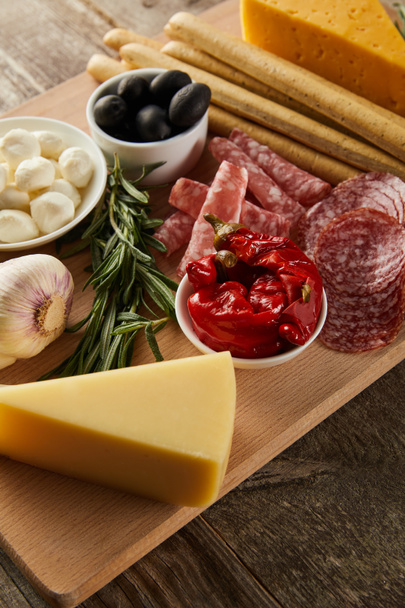 High angle view of board with breadsticks, cheese and antipasto ingredients on wooden background - Photo, image