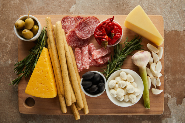 Top view of board with breadsticks, cheese, salami slices and antipasto ingredients on brown - Foto, Imagen