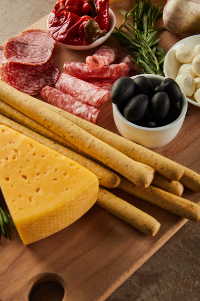 High angle view of cheese, breadsticks, salami slices, rosemary and bowls with antipasto ingredients on board on brown - Photo, Image