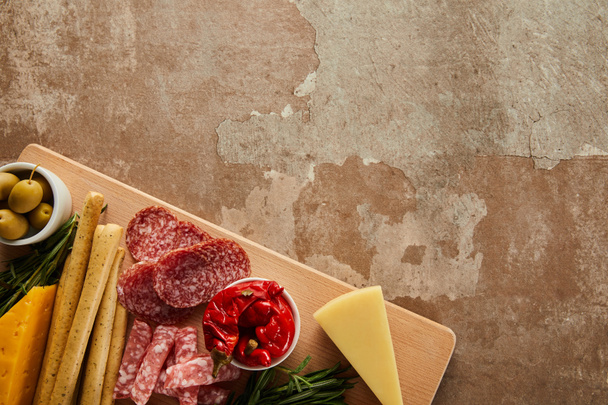 Top view of board with salami slices, cheese and antipasto ingredients on brown background - Photo, Image