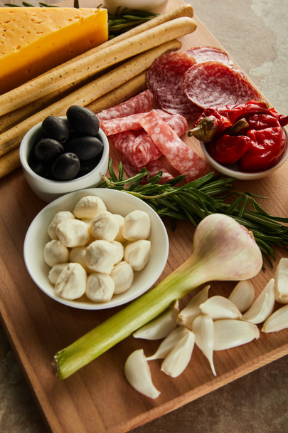 High angle view of board with antipasto ingredients and bowls with mozzarella, olives and marinated chili peppers on brown - Fotografie, Obrázek
