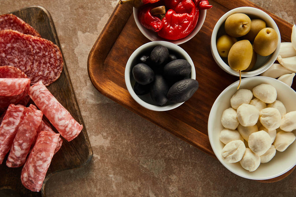 Top view of salami slices and bowls with olives, mozzarella and marinated chili peppers on boards on brown - Foto, afbeelding