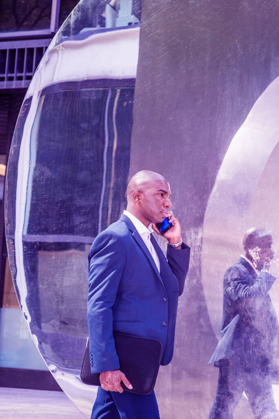 Junger afroamerikanischer Mann telefoniert, unterwegs in New York City, trägt blauen Blazer, weißes Hemd, trägt schwarze Aktentasche, geht auf die Straße, passiert Metallspiegel. Farbgefilterter Effekt - Foto, Bild