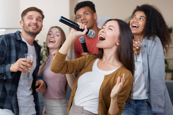 Singendes Mädchen. Junge schöne asiatische Frau hält Mikrofon und singt, während sie mit Freunden zu Hause Spaß hat. Zu Hause Karaoke spielen. Glückliche multikulturelle Menschen verbringen Zeit miteinander - Foto, Bild