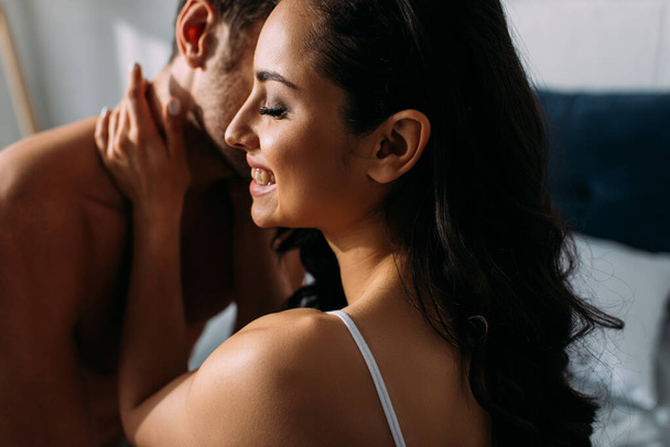 Girlfriend with closed eyes smiling and hugging boyfriend in bedroom - Fotografie, Obrázek