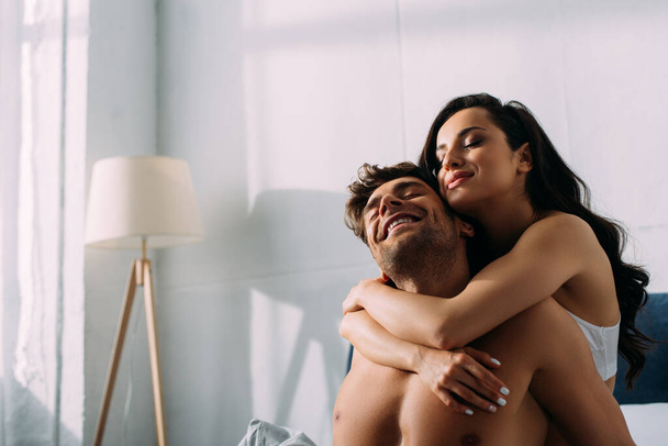 Menina feliz com os olhos fechados sorrindo e abraçando namorado por trás no quarto
 - Foto, Imagem