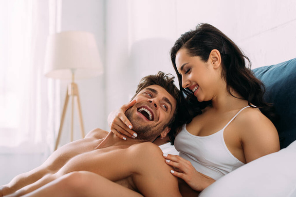 Frau lächelt und umarmt aufgeregten Mann auf Bett im Schlafzimmer - Foto, Bild