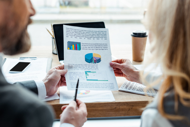 Selective focus of business people holding paper with charts near gadgets and paper cup on table  - Photo, Image
