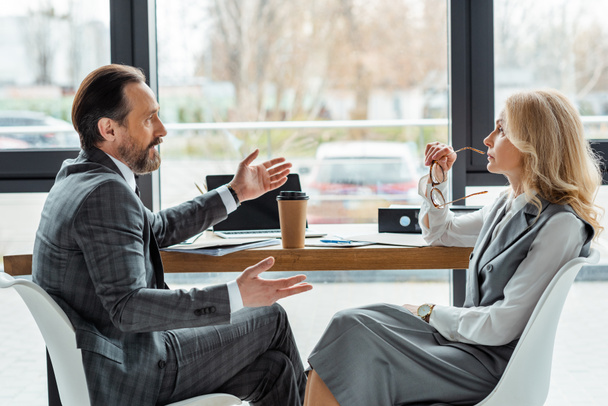 Seitenansicht eines gutaussehenden Geschäftsmannes im Gespräch mit einer Geschäftsfrau in der Nähe von Pappbechern und Dokumenten auf dem Tisch - Foto, Bild