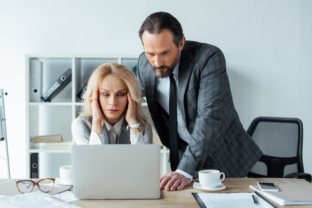 Сфокусированная деловая женщина смотрит на ноутбук рядом с бизнесменом во время работы в офисе
  - Фото, изображение