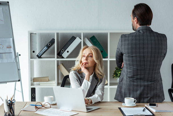Задумчивая деловая женщина смотрит в сторону ноутбука и бизнесмена в офисе
  - Фото, изображение
