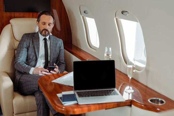 Selective focus of businessman using smartphone near laptop and champagne in plane - Photo, Image