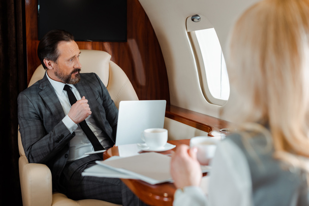 Selektiver Fokus des Geschäftsmannes mit Laptop sitzt neben Geschäftsfrau mit Tasse Kaffee im Flugzeug  - Foto, Bild