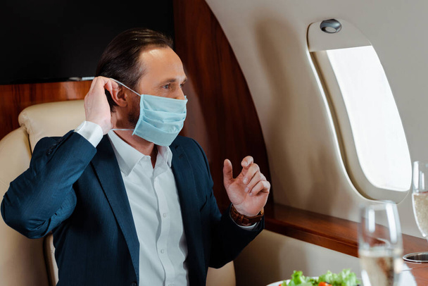 Selectieve focus van zakenman zet medische masker in de buurt van champagne en salade op tafel in het vliegtuig  - Foto, afbeelding