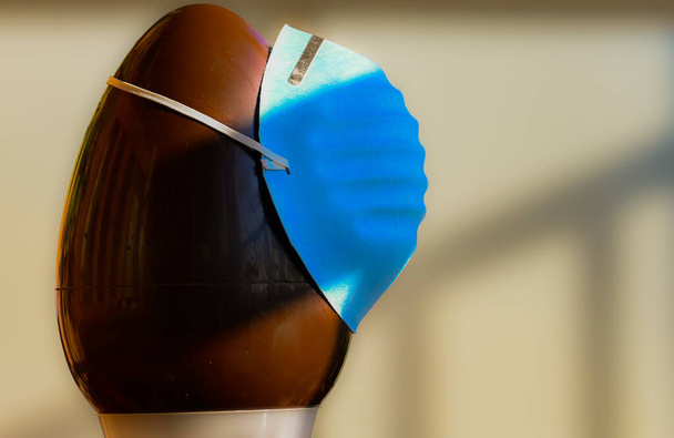 Image conceptuelle en gros plan de Pâques au moment de la pandémie de coronavirus. Un œuf en chocolat, avec une branche d'olivier à côté, porte un masque de protection bleu sur l'œuf
. - Photo, image
