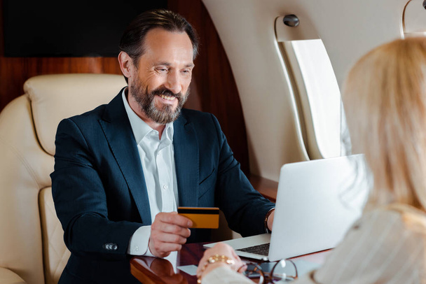 Focus selettivo di un uomo d'affari sorridente in possesso di carta di credito e utilizzando laptop vicino alla donna d'affari in aereo
  - Foto, immagini