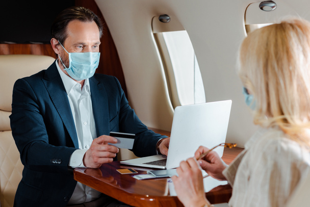 Selektiver Fokus des Geschäftsmannes in medizinischer Maske mit Kreditkarte und Laptop in der Nähe von Geschäftsfrau im Flugzeug  - Foto, Bild