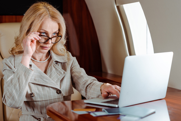 Вибіркове фокусування ділової жінки, яка тримає окуляри при використанні ноутбука біля кредитних карток і паспортів з квитками в повітрі на столі в літаку.  - Фото, зображення